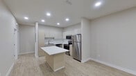 This contemporary single-floor space showcases a clean, modern design with a neutral color palette and light wood-style flooring that flows seamlessly throughout every room. The open-concept kitchen is a standout feature, outfitted with flat-front cabinetry in a warm taupe finish, a crisp white subway tile backsplash, and a full suite of stainless steel appliances including a refrigerator, electric range, and built-in microwave. A centrally positioned kitchen island with a bright white countertop provides additional workspace and serves as a natural transition into the open living area. Recessed lighting illuminates the kitchen and living spaces, creating a bright and welcoming atmosphere. The living area benefits from a large window fitted with modern day-night blinds, allowing for flexible light control, along with a front entry door that adds to the unit's accessibility. The bedroom is a calm, well-proportioned space featuring a ceiling fan with integrated lighting, a recessed light, and a window with matching day-night blinds that invite natural light. The bathroom is well-appointed with a wide dual-sink vanity, warm wood-toned cabinetry, a long wall mirror with overhead bar lighting, and a glass-enclosed shower, all finished with the same light wood-style flooring for a cohesive look throughout.