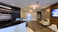 The lobby of Rutherford Station presents a modern and professional atmosphere designed to make a strong first impression. A curved reception desk with a glossy black granite top and crisp white base serves as the focal point upon entry, positioned beneath a bold navy feature wall bearing the Rutherford Station name in elegant silver lettering. Warm-toned flush-mount light fixtures complement recessed ceiling lighting throughout, casting an inviting glow across the space. Rich hardwood-style flooring extends through the main corridor, providing a seamless transition from the tiled entry area. A stainless steel elevator is centrally positioned along the back wall, offering convenient vertical access within the building. A seating area with a cushioned accent chair adds a comfortable touch for visitors. The neutral color palette of whites, grays, and deep navy tones lends the lobby a contemporary and refined aesthetic. The layout is open and well-organized, balancing functional elements such as the reception desk and elevator with welcoming design details that contribute to an overall sense of sophistication and accessibility.