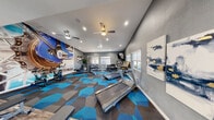 Located at 3580 E Alexander Rd in Las Vegas, NV, this fitness center is a thoughtfully designed exercise space that balances functionality with modern aesthetics. The single-room layout makes efficient use of its footprint, accommodating a diverse range of workout equipment without feeling cramped. The interior is finished in cool gray tones, with a striking large-scale bicycle chain mural dominating one wall and serving as a bold visual focal point. Vibrant blue and gray geometric patterned carpet covers the primary workout area, adding an energetic, contemporary feel to the space. A section of wood-look flooring near the entrance provides a clean transition zone. The room is well-lit through a combination of recessed ceiling lights and natural light from multiple windows fitted with horizontal blinds. Ceiling fans aid in air circulation throughout the space. Equipment includes Matrix treadmills, spin bikes, a cable strength machine, adjustable benches, a dumbbell rack, and rowing machines. A wall-mounted flat-screen television is positioned for easy viewing during cardio sessions, and a large mirror along one wall enhances the sense of depth. Tasteful wall art, potted greenery, and decorative accents complete the polished, motivating atmosphere of this fitness facility.