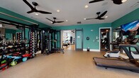 Located at 347 Arboretum Dr in Wilmington, NC, this well-appointed fitness center offers a comprehensive, multi-zone workout environment designed to accommodate a wide range of exercise needs. The facility is spread across a single floor and is divided into distinct areas, each tailored for specific types of training.

The primary workout area features bold teal accent walls, rubber flooring for safety and durability, and an array of strength training equipment, including a full dumbbell rack, power racks, barbells, benches, and cable machines. Large wall mirrors amplify the sense of space and allow users to monitor their form. Overhead, multiple black ceiling fans and recessed lighting maintain a comfortable and well-lit environment.

A dedicated cardio zone houses a row of treadmills and elliptical machines, each equipped with individual screens. Floor-to-ceiling windows along the perimeter flood this area with natural light and offer outdoor views, enhancing the overall workout experience. Large flat-screen TVs mounted on mirrored walls provide additional entertainment.

A separate spin studio, distinguished by its light gray walls and warm hardwood-style flooring, offers a focused space for cycling workouts. The room is equipped with spinning bikes, a wall-mounted display, and additional accessories including step platforms and medicine balls, making it suitable for varied group or individual sessions. Sliding barn-style doors connect the zones, providing flexible use of the overall space.