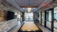 This thoughtfully designed amenity space blends industrial-chic aesthetics with modern comfort across a single, well-appointed floor. Textured brick-clad walls serve as a unifying architectural element throughout, complemented by warm wood-look flooring that adds continuity and richness to the overall design.

The centerpiece of the space is a full-size billiards table set beneath a coffered ceiling with pendant lighting. A large glass garage-style door opens toward an outdoor courtyard, flooding the area with natural light and providing a seamless indoor-outdoor connection. A wall-mounted flat-screen TV is positioned nearby for additional entertainment.

Adjacent to the game area, a lounge zone features curated seating arrangements, including leather chairs and upholstered sofas, gathered around a large-screen TV recessed into a dramatic dark accent wall with stylish sconce lighting. A built-in bookcase adds a refined touch.

The full kitchen is a standout feature, boasting dark granite countertops, dark cabinetry, a dishwasher, and a patterned tile floor. Oversized navy and gold drum pendant lights hang from a striking slatted wood ceiling, adding warmth and elegance.

A game table area with bar-height seating and a large Scrabble board wall installation offers an interactive social corner. Nearby, a small private meeting room provides an intimate setting with a round table and seating for focused gatherings. Decorative oval mirrors accent the transitional spaces throughout.