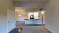 This single-floor residence showcases an open-concept layout that seamlessly connects the kitchen and living areas. The kitchen is defined by a raised peninsula with a dark countertop, offering both a functional workspace and a natural dividing point between the two zones. Stainless steel appliances, including a refrigerator and oven, add a modern touch, while a sink integrated into the peninsula and white cabinetry provide ample storage and practicality. A pendant light fixture hangs above, casting warm illumination over the space.

The living area is finished with plush wall-to-wall carpeting in a neutral tone, contributing to a comfortable and cohesive aesthetic. The entry transition features hardwood-style flooring, providing a durable and attractive threshold. Soft, warm-toned walls complement the overall palette throughout. Recessed HVAC vents are visible in the ceiling, ensuring climate control is well-integrated. Multiple doorways lead to additional rooms, suggesting a practical and functional floor plan. A wall sconce near one doorway adds supplementary lighting, enhancing the warm ambiance of the space overall.