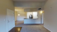 This single-floor residence showcases an open-concept layout that seamlessly connects the kitchen and living areas. The kitchen is defined by a raised peninsula with a dark countertop, offering both a functional workspace and a natural dividing point between the two zones. Stainless steel appliances, including a refrigerator and oven, add a modern touch, while a sink integrated into the peninsula and white cabinetry provide ample storage and practicality. A pendant light fixture hangs above, casting warm illumination over the space.

The living area is finished with plush wall-to-wall carpeting in a neutral tone, contributing to a comfortable and cohesive aesthetic. The entry transition features hardwood-style flooring, providing a durable and attractive threshold. Soft, warm-toned walls complement the overall palette throughout. Recessed HVAC vents are visible in the ceiling, ensuring climate control is well-integrated. Multiple doorways lead to additional rooms, suggesting a practical and functional floor plan. A wall sconce near one doorway adds supplementary lighting, enhancing the warm ambiance of the space overall.