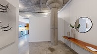 This industrial-style loft in Alameda, California showcases a striking blend of raw architectural elements and modern finishes. The open living area immediately draws attention with its soaring exposed concrete ceiling, original structural columns, and polished concrete floors — hallmarks of classic warehouse conversion design. Large, multi-pane factory-style windows flood the living space with abundant natural light while framing views of the surrounding neighborhood.

The kitchen is efficiently designed with warm wood cabinetry extending floor to ceiling, white subway tile backsplash, light countertops, and a full suite of stainless steel appliances including a built-in oven, induction cooktop, dishwasher, and over-range microwave. Track lighting above complements the industrial aesthetic throughout.

The bedroom offers a more intimate atmosphere with smooth white walls and softer lighting, providing a calm contrast to the open industrial living areas. The bathroom is cleanly appointed with large-format gray tile flooring, white subway tile surrounding the tub-shower combination, and crisp white walls. The overall design balances utilitarian character with contemporary comfort throughout.