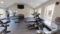 This fitness center is housed in a single-story space distinguished by vaulted ceilings that lend an open, airy quality to the interior. The architecture incorporates a painted white brick accent wall, which adds texture and visual interest, while recessed lighting and multiple large windows on opposing walls ensure the space is well-lit with natural light and views of the surrounding outdoor environment.

The flooring throughout is a durable dark rubber material, practical for a high-traffic exercise environment. A large flat-screen television is wall-mounted above the brick accent wall, providing entertainment during workouts. A full-length wall mirror is positioned strategically to maximize the sense of space and assist users with form during exercise.

The facility is comprehensively equipped with a variety of cardio machines, including multiple treadmills, elliptical trainers, a stair climber, and a stationary recumbent bike. For strength training, the space features a multi-station cable machine, a dedicated free weight area with a tiered dumbbell rack, and an adjustable weight bench. A separate connected room provides additional space and storage. The overall ambiance is clean, functional, and well-organized, offering a complete fitness environment suitable for a range of workout routines.