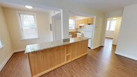 This single-story space showcases an open-concept layout that seamlessly connects the kitchen and living areas. The kitchen is outfitted with light wood cabinetry that provides ample storage, while white appliances including a refrigerator and range offer functional utility. A peninsula with a granite countertop serves as a natural divider between the kitchen and the adjoining living space, adding both style and additional workspace. The granite surface features subtle neutral toning that complements the warm wood cabinetry throughout.

Hardwood flooring runs throughout the space, lending warmth and a cohesive aesthetic across the open floor plan. The walls are finished in a soft, neutral tone that enhances the brightness of the interior. Multiple windows allow natural light to filter in, contributing to an airy and welcoming atmosphere. The entryway, featuring a classic paneled door, is conveniently accessible from the kitchen area. The overall design prioritizes an efficient, well-connected layout, making the space feel open and practical for everyday living.