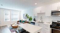 This well-designed single-floor unit offers a thoughtful layout comprising a kitchen, living room, bedroom, bathroom, and hallway. The open-concept kitchen is a standout feature, equipped with full stainless steel appliances including a gas range, built-in microwave, dishwasher, and refrigerator. White shaker-style cabinetry extends to the ceiling, complemented by a white subway tile backsplash and a undermount sink with a modern pull-down faucet. A central kitchen island with seating provides additional prep and dining space. Hardwood-style flooring flows throughout the main living areas, adding warmth and continuity.

The living room benefits from multiple windows that invite natural light, and features a bold teal accent wall that adds a distinctive pop of color against the otherwise neutral white palette. The bedroom is a comfortable, well-proportioned space with hardwood-style flooring, a window with curtains, and closet access via a hallway. The bathroom offers a clean, minimalist design with white subway tile surrounding a full bathtub and shower combination. Overall, the space presents a bright, modern aesthetic with efficient use of its compact footprint.