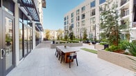 This well-appointed outdoor courtyard serves as a central amenity space within a modern multi-story residential building complex. Designed with clean, contemporary aesthetics, the courtyard is framed by a crisp white building exterior with large floor-to-ceiling glass windows and dark-framed doors that seamlessly connect the interior spaces to the outdoors. The ground level features light-toned stone pavers that extend throughout, providing a cohesive and polished surface across the entire space. A dedicated outdoor dining area is anchored by a substantial wood-top communal table surrounded by mixed seating, ideal for gatherings and al fresco meals. String lights are strung overhead, adding a warm ambiance during evening hours. Adjacent to the dining area, a compact putting green offers a recreational touch. The lounge zone is furnished with a spacious sectional sofa and accent chairs arranged around a linear fire pit, creating an inviting setting for relaxation. Raised masonry planters filled with ornamental grasses, bamboo, and trees are strategically placed throughout, introducing layers of greenery that soften the hardscape and provide a sense of privacy. The open-sky orientation allows abundant natural light to fill the courtyard throughout the day, making it a bright and welcoming outdoor retreat.