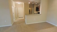 This single-floor residential unit offers a thoughtfully designed open-concept layout that maximizes both functionality and flow. The living area features soft, neutral-toned carpeting that extends throughout, creating a warm and cohesive atmosphere. Crown molding adds a refined architectural detail along the upper walls, lending a sense of polish to the interior.

The kitchen is positioned as a central focal point, defined by a peninsula-style counter with a raised breakfast bar that opens directly to the living space. Dark wood cabinetry provides rich contrast against the lighter walls and countertops, while stainless steel appliances — including a refrigerator and range — equip the space for practical everyday use. A track lighting fixture above the kitchen ensures ample task illumination.

A hallway leads from the main living area toward the entryway, which features a solid white front door and access to additional rooms. The corridor is well-lit with a ceiling-mounted light fixture and includes what appears to be a closet, adding convenient storage options. The overall color palette is consistent and calming, with warm beige tones on the walls and trim work throughout. The layout is efficient and well-organized, making excellent use of the available space on a single level.