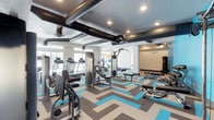 This well-appointed fitness center offers a comprehensive workout environment designed to accommodate a wide range of exercise routines. The space is anchored by a striking chevron-patterned carpet in shades of teal, gray, and beige, adding a bold visual energy that complements the room's modern aesthetic.

The cardio section lines the perimeter near large windows, housing multiple treadmills and stationary bikes that allow members to exercise in the presence of abundant natural light. At the center of the space, a multi-station cable and weight machine serves as a focal point for strength training, surrounded by adjustable benches and free weight equipment to support a variety of resistance exercises. Additional rowing and functional training stations round out the equipment offering.

The ceiling features exposed industrial-style ductwork painted in a dark finish, lending an urban loft character to the space while recessed lighting ensures consistent illumination throughout. A large flat-screen television is mounted on the wall, providing entertainment during workouts. The open layout allows for comfortable movement between stations, and the bright, clean walls contribute to an energizing atmosphere. Overall, this fitness center is a thoughtfully designed space that balances functionality with a contemporary, motivating aesthetic.