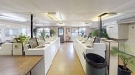 This single-floor laundromat offers a well-equipped, functional layout designed to accommodate a high volume of laundry needs. The main washing area is the centerpiece of the space, featuring two long rows of top-loading washing machines arranged back-to-back down the center of the room, providing ample capacity for simultaneous use. Natural light filters in through several windows along the front and side walls, brightening the interior alongside overhead fluorescent lighting set into a drop ceiling. The flooring throughout is a durable vinyl tile in warm earth tones.

A dedicated dryer alcove lines one wall with stacked coin-operated dryers, maximizing the use of vertical space. A coin-change machine is conveniently positioned nearby. The walls are finished with a light marbled panel treatment, giving the space a clean, utilitarian character.

A comfortable waiting area is furnished with a cushioned sofa and a long folding table lined with chairs, stocked with reading materials and decorated with a wall-mounted map. Potted plants are scattered throughout, adding a touch of greenery. A coat rack, fan, and bulletin board with posted notices round out the practical amenities available to patrons.