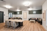 This single-floor community space offers a thoughtfully designed layout that blends function with contemporary style. The open-plan main area is anchored by a large natural wood communal table surrounded by woven accent chairs, making it ideal for group gatherings or shared work sessions. Light oak hardwood flooring runs throughout, lending warmth and visual continuity across the entire space.

A dedicated lounge alcove features a deep sectional sofa accented with colorful throw pillows, flanked by rattan side tables and a sculptural white coffee table. The walls in this area are finished with classic white board-and-batten wainscoting beneath a muted teal-gray upper wall, complemented by elegant wall sconces that create an inviting, relaxed atmosphere. Windows allow natural light to filter in, offering views of the surrounding exterior.

The kitchenette area is well-equipped with a stainless steel sink, stone countertops, cabinetry, and a dishwasher, making it practical for catered events or casual use. Framed abstract artwork decorates the walls throughout, adding a curated, gallery-like quality to the space. Access to an outdoor area is provided through a set of large glass double doors, further enhancing the connection between indoor and outdoor environments. Overall, the space presents a clean, modern aesthetic suited for community engagement and versatile programming.