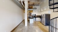Located at 1900 E Superior Ave in Cleveland's vibrant Superior district, this two-story loft unit offers a compelling blend of historic industrial character and contemporary upgrades. The main level features polished concrete tile flooring, soaring ceilings with exposed wooden beams, visible ductwork, and a striking black iron spiral staircase that serves as a focal point of the open living area. Large grid-pane windows allow natural light to fill the space. The well-appointed kitchen showcases white cabinetry, dark granite countertops, a mosaic tile backsplash, stainless steel appliances including a range, microwave, and refrigerator, and track lighting overhead. The bathroom is finished with mosaic tile accents, a tub-shower combo, a white vanity with granite countertop, and industrial-style pendant lighting. The upper level features carpeted open rooms beneath a dramatic angled ceiling of exposed timber beams and original wood planking, lending a raw, loft-like atmosphere. Additional rooms on both levels offer warm-toned hardwood flooring, pendant lighting, and ample closet storage. The overall ambiance is one of urban industrial chic, combining the building's original architectural bones with thoughtful modern renovations.