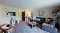 This single-floor space offers a comfortable and thoughtfully designed living environment. The living room serves as the heart of the home, featuring plush wall-to-wall carpeting that adds warmth underfoot. A dark leather sectional sofa anchors the seating area, complemented by an industrial-style coffee table on casters, decorative throw pillows, and a cozy woven throw. A wall-mounted flat-screen TV is positioned for optimal viewing, while a stylish gallery wall arrangement of framed artwork adds visual interest to the space. Ambient lighting is provided by a combination of floor lamps and accent lighting, creating a warm, inviting atmosphere throughout the room. Decorative cylindrical accent pieces and a tall floor lamp add verticality and dimension to the space. The entryway features a classic dark wood door with a decorative tile entry mat, providing a welcoming transition into the home. Neutral-toned walls throughout allow the décor and furnishings to take center stage, while the textured ceiling adds a traditional architectural detail. Overall, this space balances comfort and style in a practical, well-organized single-level layout.