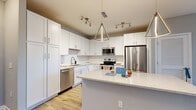 This single-floor space presents a cohesive modern design with neutral gray tones, light wood-look flooring, and white trim throughout. The kitchen serves as a standout feature, showcasing crisp white shaker-style cabinetry extending to the ceiling, a white subway tile backsplash, and a large quartz-topped island with seating potential. A full suite of stainless steel appliances—including a French door refrigerator, range, microwave, and dishwasher—adds a polished, contemporary feel. Geometric pendant lighting and track fixtures illuminate the space effectively.

The living area connects to an outdoor space via a glass door, allowing natural light to filter in. Bedrooms feature plush carpeting, ceiling fans, and generous window arrangements with blinds, ensuring both comfort and natural brightness. One bedroom offers a triple window configuration with outdoor views.

The bathrooms are finished with white shaker vanities, quartz countertops, and wood-look tile flooring. One bathroom includes a glass-enclosed shower with a window above, while the second features a clean, functional layout. Throughout, the consistent use of soft gray walls and quality finishes creates a cohesive, move-in-ready atmosphere.