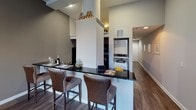 This single-floor unit offers a thoughtfully designed open layout that maximizes both functionality and style. The kitchen serves as a central hub, featuring dark granite countertops, stainless steel appliances, and a peninsula with bar-height seating for multiple guests — ideal for casual dining or entertaining. Elevated ceilings in the kitchen area add a sense of openness, and hardwood-style flooring runs throughout, lending a cohesive, modern aesthetic.

The living area is bright and inviting, benefiting from multiple large windows dressed with horizontal blinds that allow for adjustable natural light. The space is finished with warm, neutral-toned walls that complement the dark furnishings, a plush area rug, and contemporary décor including a round mirror accent and abstract wall art.

The bedroom is semi-partitioned from the main living space, providing a sense of privacy while maintaining the open feel of the floor plan. It comfortably accommodates a full-size bed and additional furnishings, with wall décor adding a personal touch.

The bathroom is clean and functional, featuring tile flooring, a full bathtub and shower combination with a striped curtain, a vanity with a light countertop, and brushed nickel fixtures and grab bars. The space is practical and well-maintained throughout.