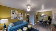 This Sundance one-bedroom, one-bathroom residence is a single-floor unit designed with a functional and stylish open-concept layout. The living area serves as the heart of the home, featuring warm wood-style flooring, a textured ceiling, and a central light fixture that provides ample illumination. Bold accent walls in a muted gold-green tone add a distinctive design element throughout, complemented by tasteful wall art that enhances the contemporary aesthetic.

The living space flows seamlessly into the dining area, which is illuminated by an elegant pendant chandelier and offers room for a full dining set — ideal for everyday meals or entertaining guests. Adjacent to the dining area, the kitchen is efficiently laid out with white cabinetry, dark appliances including a refrigerator and range, and a practical design that maximizes storage and workspace.

The open sightlines between the living, dining, and kitchen areas create a sense of spaciousness and connectivity throughout the unit. Consistent wood-style flooring ties the common areas together, while neutral gray trim and baseboards provide a clean, polished finish. This unit offers a well-balanced combination of comfort, style, and practicality suited for everyday living.