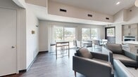 Located at 5200 W 98th St in Bloomington, MN, this single-floor combined kitchen and living room space offers a thoughtfully designed, open-concept layout with contemporary finishes throughout. The kitchen is well-appointed with gray shaker-style cabinetry, sleek quartz countertops with subtle veining, a stainless steel refrigerator, a built-in wall oven, and a black matte faucet over an undermount sink. A large quartz-topped island with upholstered bar seating serves as both a functional workspace and a casual dining area. A horizontal pass-through window connects the kitchen to the adjacent space, adding an architectural detail that enhances openness. The living area features warm hardwood-style flooring, soaring ceilings, and a wall-mounted electric fireplace flanked by modern seating. A flat-screen television is mounted on the wall, and a sleek track light with a security camera is positioned above the entrance. Large sliding glass doors along one wall flood the space with generous natural light and offer views of the surrounding outdoor landscape. The neutral color palette of whites and soft grays creates a clean, inviting ambiance throughout.
