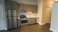 This single-floor space presents a clean and contemporary design that emphasizes both functionality and style. The kitchen serves as a central highlight, outfitted with a full suite of stainless steel appliances including a gas range with oven, over-the-range microwave, refrigerator, and dishwasher. Flat-front cabinetry in a warm taupe-gray finish lines the walls, offering generous storage while contributing to the modern aesthetic. A classic subway-style stone tile backsplash adds texture and visual interest behind the countertops, complementing the light-toned counter surfaces. The undermount sink is paired with a sleek modern faucet, adding a refined touch to the space. Rich, dark hardwood-style flooring runs throughout, providing a warm contrast to the lighter cabinetry and walls. The space benefits from a clean, open layout that allows for easy movement and a sense of openness. Smooth walls and crisp white baseboards lend a polished, well-maintained appearance throughout. Overall, the space strikes a balance between modern urban design and practical livability, making it an inviting and highly functional environment.