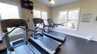 This fitness center offers a bright and functional environment designed to support an active lifestyle. The space is outfitted with multiple Life Fitness treadmills and additional cardio equipment, all arranged to maximize usability and comfort. Large windows along the walls flood the room with natural light and provide pleasant views of the surrounding greenery, creating an inviting atmosphere that makes workouts more enjoyable. The flooring features durable, dark rubber matting suited for high-traffic exercise use. A wall-mounted flat-screen television is positioned for easy viewing from the cardio machines, offering entertainment during workouts. Overhead lighting supplements the natural light, ensuring the space remains well-lit at all times. The neutral-toned walls and clean, uncluttered layout contribute to a focused and motivating environment. The room is thoughtfully designed to accommodate multiple users simultaneously while maintaining a sense of openness. Overall, this fitness center combines practical amenities with a comfortable, light-filled setting to deliver a well-rounded exercise facility.