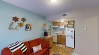 This single-floor, one-bedroom, one-bathroom unit offers a practical and charming layout that maximizes its compact footprint. The living area and kitchen flow seamlessly together in an open-concept arrangement, creating a connected and functional living environment. The living space features soft blue-green accent walls that lend a calm, welcoming ambiance, complemented by warm hardwood-style flooring that runs throughout the unit.

The kitchen is efficiently designed with warm wood cabinetry providing ample storage, white appliances including a refrigerator, range, and microwave, and a dedicated under-cabinet ventilation system. Open shelving adds both display and storage opportunities, while the layout keeps essentials within easy reach.

The living area accommodates comfortable seating and personal touches, with wall décor adding character and personality to the space. Ceiling-mounted light fixtures provide consistent illumination throughout, and overhead HVAC vents ensure climate control. The overall ambiance is cozy and lived-in, making this unit a practical choice for those seeking a well-utilized, single-floor living space.