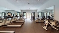 Located at 2005 Treehills Pkwy in Stone Mountain, Georgia, this fitness center offers a spacious, single-floor workout facility designed to support a comprehensive exercise routine. The gym features rubber flooring throughout, providing a durable and comfortable surface suited for a variety of fitness activities. Large wall-to-wall mirrors line the sides of the room, enhancing the sense of openness while allowing users to monitor their form during workouts. Recessed lighting illuminates the space evenly, complemented by natural light streaming in through multiple windows and a set of French doors that open to an outdoor area, offering views of surrounding greenery. The cardio section is well-appointed with a row of TRUE-brand treadmills and elliptical machines, alongside upright and recumbent stationary bikes. A multi-station strength training cable machine occupies the center of the room, providing options for a range of resistance exercises. Additional strength equipment, including a vertical knee raise station and free-weight benches, rounds out the available amenities. The layout is open and uncluttered, allowing for easy movement between equipment zones. A textured ceiling and neutral white walls contribute to a clean, functional aesthetic throughout the facility.
