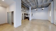 This single-floor space showcases a striking industrial-modern aesthetic, combining raw architectural elements with contemporary finishes. The open-concept layout is anchored by polished concrete floors that extend throughout, reflecting light and contributing to the expansive feel of the interior. Soaring ceilings reveal exposed structural beams, metal ductwork, and conduit, lending an authentic loft character to the space. Pendant lighting fixtures hang at varying heights, providing both functional illumination and a design focal point against the crisp white walls and ceiling.

Structural columns are integrated thoughtfully into the floor plan, defining zones without interrupting the overall sense of openness. The kitchen area features dark cabinetry, a stainless steel refrigerator, and a white countertop peninsula that serves as a natural divider between the cooking and living areas. Built-in open shelving adds practical storage near the kitchen zone.

The generous main living area offers a versatile, unobstructed footprint ideal for a variety of configurations. A closet or utility area is visible toward the back of the space, adding functional storage. Overall, the space balances an urban industrial atmosphere with clean, modern finishes, making it a highly adaptable and visually compelling environment.
