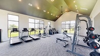 This modern fitness center in Huntsville, Alabama offers a spacious and well-appointed workout environment designed to support a full range of exercise routines. The space is defined by its striking vaulted ceiling, finished in a bold dark tone that creates a dramatic architectural contrast against the crisp white walls. Recessed lighting and stylish pendant fixtures illuminate the room evenly, ensuring a bright and energizing atmosphere throughout the day.

Large, grid-paned windows line multiple walls, flooding the interior with abundant natural light and offering views of the surrounding green landscape — an inviting backdrop for any workout. The floor is covered in durable, textured carpet tile, adding both comfort and a polished, contemporary aesthetic.

The cardio zone is well-stocked with treadmills, elliptical machines, a stair climber, and stationary bikes, all positioned to take advantage of the outdoor views. The strength training area features a multi-station cable machine, a power rack with free weights, adjustable benches, and additional resistance equipment. Full-length mirrors on one wall enhance the sense of space and support proper form during lifts.

Overall, the fitness center strikes a balance between functional utility and modern design, providing a motivating and comfortable environment for residents in Huntsville, AL.