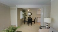 This single-floor residence offers an inviting, open-concept layout designed for comfortable everyday living. The dining area serves as a central gathering point, featuring plush carpeting underfoot and warm, neutral-toned walls that create a welcoming atmosphere. A ceiling fan overhead provides both function and a subtle design element, while a round wall mirror adds a decorative focal point to the space. The dining area flows seamlessly into the adjacent kitchen, which is visible through a wide, open passage framed by a structural beam. The kitchen is finished with white cabinetry offering ample storage, light-toned countertops, and tile flooring that contrasts neatly with the carpeted dining and living areas. A window in the dining area allows natural light to filter into the space, brightening the interior and enhancing the airy feel. Lush indoor plants are incorporated throughout, adding a touch of greenery and warmth to the neutral palette. The living area, partially visible at the foreground, continues the cohesive design language with coordinating tones and furnishings. The textured ceiling and clean architectural lines complete the well-maintained, functional character of this single-level home.