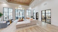 Located at 4415 Percival Rd in Columbia, SC, this single-floor community space offers a thoughtfully designed, open-concept layout that combines a living room and kitchen area into one cohesive, welcoming environment. The space immediately impresses with its soaring ceilings, wood-beam ceiling detail, and a dramatic floor-to-ceiling arched window wall with black steel grid framing that floods the interior with natural light and provides views of the surrounding community.

The main living area features light wood-look tile flooring throughout, anchored by a large, modular cream sectional sofa beneath an elegant circular chandelier. Multiple seating zones create comfortable gathering spaces, while crisp white walls and black-framed doors and windows lend a clean, modern aesthetic.

The kitchen nook is a standout feature, showcasing warm wood shiplap accents, gray cabinetry with white countertops, and teal pendant lighting that adds a playful pop of color. Built-in open wood shelving lines the adjacent walls, styled with books and greenery against a patterned floral wallcovering, adding warmth and personality.

A gallery wall displaying curated artwork alongside a wall-mounted display screen adds a functional yet artistic touch. Additional access doors open directly to outdoor areas, enhancing the flow between interior and exterior. The overall ambiance is bright, airy, and modern, designed to foster community connection.