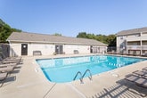 Located at 1272 Union Rd. in Gastonia, North Carolina, this community amenity space offers residents two key recreational facilities designed for both fitness and relaxation. The outdoor pool area is a standout feature, showcasing a generously sized swimming pool with clear turquoise water, clearly marked depth indicators, and a no-diving designation for safety. A spacious concrete pool deck surrounds the water, furnished with multiple lounge chairs and patio seating, perfect for enjoying the Carolina sunshine. The entire pool area is enclosed by a secure iron fence, providing privacy and safety, with the residential building's balconies visible in the background. The fitness center is thoughtfully designed with a two-tone paint scheme — warm gray on the lower walls accented by a bold teal stripe near the ceiling — giving the space a modern, energetic feel. Rubber flooring throughout provides comfort and durability underfoot. The room is equipped with a multi-function cable weight machine, a row of elliptical trainers, treadmills, and additional cardio equipment. Wall-mounted mirrors enhance the sense of space, while windows allow natural light to filter in. Posted exercise rules and a fire safety station reflect a well-maintained, safety-conscious environment.