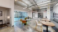 This single-floor commercial space delivers a compelling industrial-chic aesthetic rooted in raw architectural character. Polished concrete floors stretch throughout, complemented by soaring exposed ceilings where structural beams, ductwork, and conduit are left fully visible — a deliberate design choice that adds an authentic, loft-like energy to the environment.

The main open area serves as a versatile collaborative hub, furnished with an eclectic mix of round tables, modern chairs in warm wood tones with green metal frames, and plush mustard-yellow lounge seating arranged around low coffee tables. A dramatic large-format black-and-white photographic mural anchors one wall, adding historical depth and visual impact. Pendant Edison bulbs hang in clusters, providing warm ambient lighting alongside industrial fluorescent strips overhead.

A corridor with glass-paneled doors connects to additional areas, including a quieter lounge zone with exposed raw-concrete walls and intimate seating. A private office offers a focused work environment, neatly appointed with a dark desk, task chair, and curated wall art. Large glass sliding doors along one wall flood the space with natural light and provide direct access to an exterior area, enhancing the open, airy feel. The overall ambiance balances gritty industrial elements with thoughtfully curated modern furnishings, creating a dynamic and functional workspace.