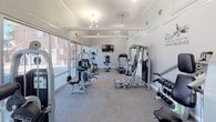 This fitness center is a thoughtfully designed, single-floor exercise facility that offers a bright, open, and motivating environment for residents. The space is bathed in natural light, thanks to an expansive wall of floor-to-ceiling windows running along one side of the room, providing views of the outdoor pool area and surrounding greenery. The light-colored walls and neutral carpet flooring keep the atmosphere clean and energizing.

The facility is divided into two functional zones. The cardio area is equipped with a treadmill, elliptical trainer, and stationary bike, all positioned beneath a wall-mounted flat-screen television — ideal for staying entertained during workouts. Adjacent to this, the strength training area features multiple cable and resistance machines, a multi-purpose weight rack, and a dumbbell station, offering a comprehensive range of equipment for full-body training.

Overhead, a combination of pendant lights and track lighting ensures the space is well-lit throughout. Motivational wall decals — featuring phrases like "Beast Mode" and "Will It Be Easy? Nope. Worth It? Absolutely." — add character and inspiration to the space. The layout provides ample room to move between equipment comfortably, making it a practical and welcoming fitness destination.