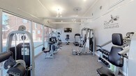 This fitness center is a thoughtfully designed, single-floor exercise facility that offers a bright, open, and motivating environment for residents. The space is bathed in natural light, thanks to an expansive wall of floor-to-ceiling windows running along one side of the room, providing views of the outdoor pool area and surrounding greenery. The light-colored walls and neutral carpet flooring keep the atmosphere clean and energizing.

The facility is divided into two functional zones. The cardio area is equipped with a treadmill, elliptical trainer, and stationary bike, all positioned beneath a wall-mounted flat-screen television — ideal for staying entertained during workouts. Adjacent to this, the strength training area features multiple cable and resistance machines, a multi-purpose weight rack, and a dumbbell station, offering a comprehensive range of equipment for full-body training.

Overhead, a combination of pendant lights and track lighting ensures the space is well-lit throughout. Motivational wall decals — featuring phrases like "Beast Mode" and "Will It Be Easy? Nope. Worth It? Absolutely." — add character and inspiration to the space. The layout provides ample room to move between equipment comfortably, making it a practical and welcoming fitness destination.