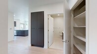 This single-floor residence showcases a clean, contemporary design with light hardwood flooring and crisp white walls throughout. The open-concept kitchen serves as a central hub, featuring a large dark-finished island with an integrated sink, flat-panel dark cabinetry, white quartz countertops, a white tile backsplash, and stainless steel appliances including a refrigerator, range, and microwave. Multiple windows and a glass-panel entry door allow natural light to fill the kitchen and adjacent living areas.

The living and bedroom spaces are generously proportioned, benefiting from oversized windows that frame lush, tree-lined outdoor views and invite abundant daylight. Recessed lighting supplements natural illumination throughout.

A hallway connects the main living areas to the bathroom and utility zones. The bathroom is finished with a wide, white quartz double-vanity counter, flat-panel cabinetry, an illuminated mirror, and clean tile flooring. A separate water closet and grab bars indicate accessible design. The hallway also features a tall, dark flat-panel storage cabinet and built-in shelving. An in-unit stacked washer and dryer is tucked neatly within a dedicated alcove, adding practical convenience to this well-appointed residence.