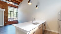 This distinctive two-floor space masterfully combines industrial character with contemporary design. The architecture showcases exposed brick walls, warm wood-plank ceiling beams, and visible ductwork, all hallmarks of adaptive reuse construction that lend the space a rich, textured aesthetic.

The open-concept living area features light wood-tone luxury vinyl plank flooring and soaring ceilings that amplify the sense of volume throughout. Large steel-framed windows with brick arch detailing flood the interior with natural light while offering views of surrounding greenery.

The kitchen is a standout feature, centered around a generous island with crisp white quartz countertops, an undermount stainless steel sink, and warm natural wood cabinetry. Matte black pendant lights hang overhead, adding a modern industrial accent. The kitchen is fully equipped with stainless steel appliances including a refrigerator, range, microwave, and dishwasher.

Additional rooms include well-proportioned bedrooms with closet storage and wood ceiling details, and a clean, modern bathroom featuring a wood-toned vanity, white quartz countertop, and wood-look tile flooring. A second floor extends the living space further, offering versatile use. Throughout, the palette of neutral tones, natural wood, and exposed masonry creates a cohesive, contemporary-industrial ambiance.