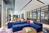 Located at 501 NW Seventh Ave in Fort Lauderdale, FL, this single-floor amenity space offers a generously sized, open-plan layout designed for socializing, relaxing, and recreation. The interior is defined by a contemporary aesthetic that blends clean architectural lines with warm, eclectic furnishings. Light hardwood flooring runs throughout, complemented by vibrant, layered Persian-style area rugs that anchor distinct seating zones.

Dramatic floor-to-ceiling windows and glass doors line the perimeter, flooding the space with natural light while framing lush views of tropical palms, landscaped gardens, and a pool area beyond. Bold navy blue modular sofas, cognac leather lounge chairs, and bistro-style café tables create a variety of comfortable gathering spots suited to different activities and group sizes.

Striking steel-framed glass partition walls divide the space into semi-private rooms, including what appears to be a game room featuring a foosball table and colorful bowling balls set against an exposed brick accent wall. A library nook with built-in illuminated shelving displays books and art objects, providing a quieter retreat. Recessed lighting, teal ceiling accents, and curated artwork contribute to a polished yet inviting atmosphere throughout this well-appointed amenity floor.