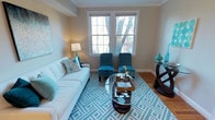 This single-floor space showcases a thoughtfully designed living area that blends comfort with contemporary style. The room is anchored by warm hardwood flooring and neutral beige walls, creating a versatile backdrop for its vibrant teal and white color scheme. Two large double-hung windows along the far wall flood the space with natural light and offer views of a tree-lined outdoor setting, enhancing the room's open and airy feel. Classic white crown molding and clean baseboards frame the space with a refined architectural touch. The living area is furnished with a tufted white sofa accented by teal throw pillows, complemented by a pair of plush teal accent chairs arranged symmetrically near the windows. A round glass-top coffee table with a dark sculptural base serves as a central focal point, resting atop a bold geometric teal and white area rug that ties the color palette together. A dark wood console table with decorative teal accessories is positioned along one wall, adding depth and visual balance. A softly lit table lamp contributes to the warm, welcoming ambiance of the space.
