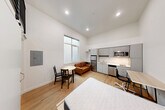 Located along University Way NE in Seattle's vibrant University District, this efficiently designed studio unit makes excellent use of its compact footprint. The main living area is open-plan, seamlessly combining sleeping, dining, and living functions under soaring ceilings that lend a sense of airiness well beyond the unit's modest size. Light wood-tone flooring runs throughout, complementing the crisp white walls and recessed lighting overhead.

The kitchen is thoughtfully outfitted with grey flat-front cabinetry extending to the ceiling, stainless steel appliances including a refrigerator, microwave, and dishwasher, a single-basin sink, and a built-in countertop workspace that doubles as a desk — ideal for working or studying from home. A small dining table and seating area are positioned near the window, which allows natural light to filter into the space.

A dedicated laundry area houses a full-size washer and dryer with overhead shelving, providing practical convenience. The bathroom features a clean, accessible layout with grab bars, a toilet, vanity, and tile flooring. A loft-style metal railing above the entry adds an industrial design accent. This unit is well-suited for students or individuals seeking a functional, modern living space in a prime Seattle neighborhood.