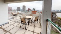 Located at 100 Memorial Dr in Cambridge, MA, this elevated outdoor space offers a remarkable vantage point overlooking the Charles River and the iconic Boston skyline. The space is comprised of a single-level terrace and rooftop deck, both finished with large-format tile flooring in a neutral palette that complements the open, airy environment.

The covered terrace features a classic French door entry with warm wood-toned frames, flush ceiling-mounted lighting, and a glass-railed balustrade that preserves unobstructed sightlines to the surrounding cityscape. Outdoor seating with tables and chairs is arranged to take full advantage of the panoramic views, creating an ideal setting for gatherings or quiet enjoyment.

An adjacent open-air rooftop deck expands the outdoor living experience further, with additional seating clusters set against a backdrop of Cambridge and Boston's urban skyline. From this elevated position, sweeping views of the Charles River, Memorial Drive, and the Boston Back Bay skyline are prominently on display, with landmarks and waterfront greenery visible in the distance.

A long wraparound balcony runs along the building's perimeter, offering continuous views of the neighboring residential buildings and the broader Cambridge streetscape. Together, these outdoor areas form a compelling and well-positioned elevated retreat in the heart of Cambridge.