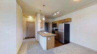 This single-floor residence offers a thoughtfully designed open-concept layout that seamlessly connects the kitchen and living areas. The kitchen is a functional centerpiece, featuring warm honey-toned wood cabinetry, granite-look countertops, and a full suite of appliances including a stainless steel refrigerator, black range, and built-in microwave. A peninsula breakfast bar with pendant lighting overhead adds both style and casual dining convenience. Track lighting illuminates the kitchen workspace, while the transition between hardwood-style flooring in the kitchen and plush carpeting in the living area creates distinct yet connected zones. The entryway, visible from the kitchen, features a solid paneled door and offers a welcoming first impression. Neutral wall tones and light-colored ceilings throughout lend the space a clean, airy ambiance. The open sightlines from the kitchen to the adjacent living area enhance the overall sense of spaciousness. This residence balances practical design with comfortable living, making it an ideal space for both everyday routines and relaxed gatherings.