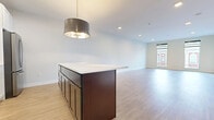 This single-floor space showcases a contemporary design with a thoughtfully arranged open layout. The kitchen is a standout feature, equipped with stainless steel appliances including a range, microwave, and refrigerator, complemented by white upper cabinetry and a patterned tile backsplash. A substantial dark-wood island with a bright white countertop anchors the kitchen, illuminated by a modern drum pendant light. The kitchen flows seamlessly into the living area, which is finished with light wood-style flooring throughout. Tall, slim windows with black frames line the living space, allowing generous natural light and offering views of the surrounding urban streetscape.

The bedroom features neutral-toned carpet and crisp white walls, creating a calm and functional space. Exposed ductwork adds an industrial touch to the bedroom corridor. The bathroom is well-appointed with a glass-enclosed walk-in shower featuring textured tile walls, a built-in shelf, and a clean white tile floor. Recessed lighting throughout the unit, combined with bright white walls and light flooring, contributes to an airy, modern ambiance across all areas of the space.