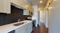 This single-floor unit showcases a cohesive modern design with a thoughtful layout and quality finishes throughout. The kitchen is a standout feature, equipped with light wood-grain cabinetry, stainless steel appliances including a French door refrigerator, electric range, built-in microwave, and dishwasher. A bold dark subway tile backsplash contrasts with white countertops, adding a contemporary edge. Recessed lighting and dark hardwood-style flooring complete the space.

The open living area flows directly from the kitchen, benefiting from large windows with horizontal blinds that invite natural light and provide views of surrounding greenery. A sliding glass door offers additional outdoor access. The bedroom features soft carpet flooring, neutral-toned walls, and a recessed ceiling light, providing a calm and practical retreat.

A dedicated hallway leads to a bathroom featuring a wood-grain vanity with a white countertop, a wall mirror, and dark hardwood-style flooring. Nearby, a spacious walk-in closet with wire shelving and a stacked washer and dryer unit offer excellent storage and in-unit laundry convenience. Sliding barn-style doors add a functional, modern touch to the interior corridors.