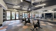 This 590-square-foot fitness center is a single-floor facility thoughtfully designed to accommodate a full range of workout options. The space features a striking hexagonal tile floor in a blend of gray, tan, and copper tones, adding a modern, dynamic aesthetic throughout. Multiple spin bikes are arranged across the open floor plan, making the room well-suited for cycling workouts. A dedicated strength training area includes a multi-station cable machine, a dumbbell rack with a full set of free weights, and medicine balls, supported by floor-to-ceiling mirrors along one wall that enhance the sense of space and allow users to monitor their form. A large flat-screen television is mounted on the wall, providing entertainment during workouts. The dark, textured ceiling features recessed linear LED lighting and multiple ceiling fans, ensuring the space remains well-lit and well-ventilated. Floor-to-ceiling glass doors and large windows along one wall flood the interior with natural light and offer views of an outdoor pool area, creating an inviting and energizing atmosphere. The overall design combines functionality with a sleek, modern aesthetic suitable for a residential amenity fitness center.