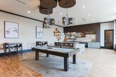 Located at 2–14 S Clayton St in Lawrenceville, GA, this single-floor amenity space offers a sophisticated and inviting environment designed for socializing and recreation. The interior showcases a cohesive modern aesthetic anchored by warm wood-plank flooring, shiplap-style wood accent walls, and carefully layered lighting throughout.

The lounge area serves as the focal point of the space, featuring a tufted sofa, curated side tables, and a large-scale abstract artwork mounted against a full wood-paneled accent wall illuminated by a row of recessed spotlights. A large flat-screen display adds a media component ideal for communal viewing.

Adjacent to the lounge, a billiards table anchors the central gathering zone, providing a natural hub for social activity. Pendant lighting with Edison-style bulbs hangs above the dining and bar areas, contributing to the warm, ambient atmosphere.

The kitchen and bar area is a standout feature, equipped with sleek gray cabinetry, stainless steel appliances, a beverage refrigerator, a mosaic tile backsplash, and an expansive concrete-finish island with bar seating. Decorative sculptural wood art pieces adorn the walls, adding an organic, artistic contrast to the contemporary finishes. Large windows allow natural light to fill the kitchen zone, offering views of the surrounding Lawrenceville streetscape.