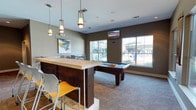 This single-floor amenity lounge is thoughtfully designed to serve as a social and recreational hub. The space is anchored by a dark wood pool table positioned at the center, providing a focal point for leisure activity. Adjacent to it, a substantial bar-height counter with a light wood top offers casual seating with modern, colorful stools in white and orange, adding a vibrant contrast to the neutral palette.

Pendant lights with warm-toned fixtures hang above the bar area, complementing the recessed lighting throughout the room to create a layered, inviting atmosphere. The walls are finished in a warm taupe tone, grounded by carpeted flooring that adds comfort underfoot.

Large windows along the far wall flood the interior with natural light and provide views of the outdoor surroundings, enhancing the open, airy feel of the space. Colorful artwork adorns the walls, contributing energy and personality to the environment. A wall-mounted flat-screen television is positioned for easy viewing across the room. A secondary seating area with chairs and a table is visible near the windows, offering a quieter spot for conversation or casual gatherings.