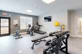 Located at 2944 Landrum Dr SW in Atlanta, GA, this fitness center offers a thoughtfully designed exercise space ideal for a comprehensive workout routine. The room features a clean, modern aesthetic with a two-tone color scheme of light gray and charcoal accent walls, complemented by durable speckled flooring suited for high-traffic athletic use. Large panel light fixtures mounted on the ceiling provide bright, even illumination throughout, while multiple windows along the walls allow ample natural light and views of the surrounding greenery, creating an open and energizing atmosphere. The space is well-equipped with a variety of fitness equipment, including a treadmill, a stationary spin bike, and a recumbent exercise bike for cardiovascular training. A multi-tiered dumbbell rack stocked with a full range of free weights, a flat workout bench, and a medicine ball storage rack with assorted exercise balls support strength and functional fitness training. A wall-mounted flat-screen television adds convenience for entertainment during workouts. A large floor-to-ceiling mirror on one wall enhances the sense of space and allows users to monitor their form. The room is finished with clean white baseboards and smooth walls, contributing to an overall polished and professional environment.