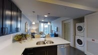 This contemporary single-floor residence showcases a thoughtfully designed open-concept layout that integrates the kitchen and living areas into one cohesive, functional space. The kitchen serves as the centerpiece, featuring a large island with a white quartz countertop and an undermount double-basin stainless steel sink. Dark cabinetry provides a striking contrast to the light countertops, while stainless steel appliances, including a dishwasher, add a polished, modern finish. Pendant lighting hangs above the island, enhancing both ambiance and task lighting. The living area beyond is bright and inviting, benefiting from large windows that allow natural light to fill the room and offer views of the surrounding cityscape. A wall-mounted flat-screen television and carefully chosen furnishings complete the lounge space, creating a comfortable and stylish environment. The ceiling features a recessed tray detail with a decorative finish, adding architectural interest overhead. A stacked washer and dryer unit is conveniently tucked into a dedicated nook, offering in-unit laundry. The overall design balances modern aesthetics with practical functionality throughout.