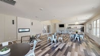The community clubhouse at Villas of Jeffersonville is a thoughtfully designed single-floor gathering space that blends modern aesthetics with functional amenities. Throughout the entire space, a striking multi-toned wood-look plank flooring ties the areas together, featuring varying shades of gray, brown, and blonde tones arranged in a dynamic pattern that adds visual depth and texture underfoot.

The main gathering area offers generous open space with multiple dining tables and modern white metal chairs, illuminated by elegant pendant chandeliers. Large windows along the perimeter flood the room with natural light while providing views of the surrounding community. A mounted flat-screen television serves as a focal point for communal viewing.

A cozy lounge nook features a sleek, linear gas fireplace set within a white tile accent wall, accompanied by comfortable seating and a patterned area rug, creating an intimate retreat within the larger space. Framed artwork and curated décor enhance the refined atmosphere throughout.

The game room extension houses a full-size pool table beneath a stylish chandelier, with multiple windows ensuring a bright and inviting environment. A kitchenette area with bar seating and cabinetry rounds out the amenities, offering a convenient space for entertaining. Sliding glass doors provide direct access to an outdoor area, seamlessly connecting indoor and outdoor living.