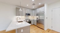 Located at 1435 51st Street in North Bergen, NJ, this 2-bedroom, 1-bathroom unit offers a well-designed open-concept layout with a clean, modern aesthetic. Light hardwood flooring flows throughout the entire unit, creating a cohesive and bright atmosphere.

The kitchen is a standout feature, equipped with grey shaker-style cabinetry, a striking marble-veined quartz waterfall island with an undermount sink, a textured tile backsplash, and a full suite of stainless steel appliances including a refrigerator with ice maker, range, microwave, and dishwasher. Recessed lighting and a pendant light over the island complete the polished look. The kitchen opens directly into the living area, which benefits from natural light through a window overlooking lush greenery.

Both bedrooms are bright and serene, featuring hardwood floors, white walls, and windows with tree-lined views. The primary bedroom offers a comfortable footprint with ample wall space.

The bathroom features large-format cream floor tiles, a grey vanity with a white countertop, a framed mirror, and an eye-catching mosaic tile accent wall in warm amber and brown tones.

The unit also includes a hallway connecting the main living space to the bedrooms, providing a practical and organized flow throughout.